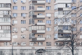 An apartment building with a beige facade features multiple windows, some with balconies enclosed by glass. Air conditioning units are mounted beneath several windows. The lower level has commercial establishments, including a visible post office sign. Leafless trees partially obscure parts of the building.