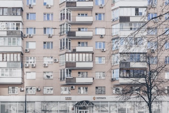 An apartment building with a beige facade features multiple windows, some with balconies enclosed by glass. Air conditioning units are mounted beneath several windows. The lower level has commercial establishments, including a visible post office sign. Leafless trees partially obscure parts of the building.