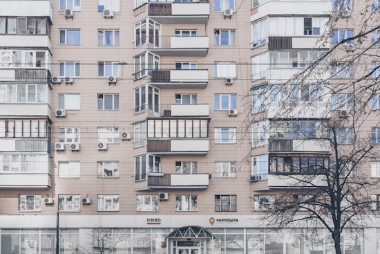 An apartment building with a beige facade features multiple windows, some with balconies enclosed by glass. Air conditioning units are mounted beneath several windows. The lower level has commercial establishments, including a visible post office sign. Leafless trees partially obscure parts of the building.