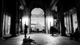 A black and white scene featuring a violinist playing under an ornate arched gateway. Tall columns frame the scene, and a distant view of the Louvre Pyramid is visible through a gated fence. Several people are present, creating a lively yet serene atmosphere.
