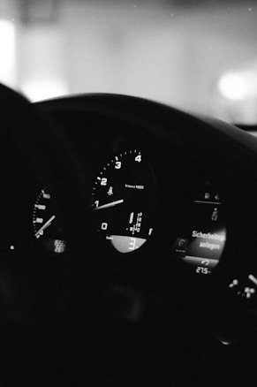 A black and white image capturing the dashboard of a vehicle, featuring visible speedometer and tachometer gauges. The tachometer needle is pointing between 1 and 2, while various digital displays show information such as gear position and temperature. The interior is dimly lit, conveying a moody atmosphere.