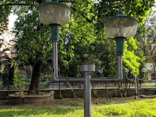 A pair of vintage-style lamps are mounted on a metal post in a park setting. Surrounding the lamps are lush green trees and foliage, with some buildings visible in the background. The ground is covered with grass, and there is a circular concrete planter near the base of a large tree.