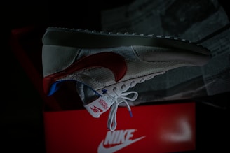 A single sneaker is prominently displayed against a dark background. The shoe features a red swoosh, white mesh, and suede material with blue and red accents. It is positioned over an open red shoe box with visible branding. Newspapers are partially visible in the background.