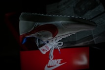 A single sneaker is prominently displayed against a dark background. The shoe features a red swoosh, white mesh, and suede material with blue and red accents. It is positioned over an open red shoe box with visible branding. Newspapers are partially visible in the background.