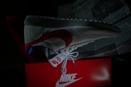 A single sneaker is prominently displayed against a dark background. The shoe features a red swoosh, white mesh, and suede material with blue and red accents. It is positioned over an open red shoe box with visible branding. Newspapers are partially visible in the background.