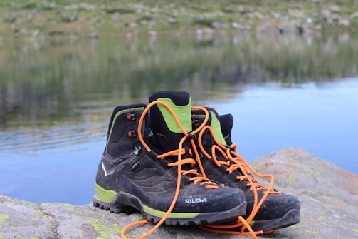 A pair of black hiking boots with orange laces and green accents are placed on a large rock beside a calm body of water. In the background, there is a blurred view of grass and rocks, creating a serene outdoor setting.