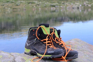 A pair of black hiking boots with orange laces and green accents are placed on a large rock beside a calm body of water. In the background, there is a blurred view of grass and rocks, creating a serene outdoor setting.