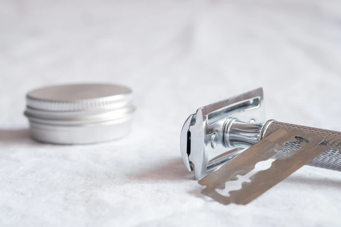 A silver safety razor with the blade exposed beside a small round tin on white cloth (stand-in for a dermaplaning blade)