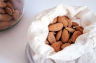A white cloth bag filled with whole almonds, with a glass jar of almonds partially visible in the background. The almonds have a natural brown hue and an oval shape, and the cloth bag is casually open, showcasing the nuts.