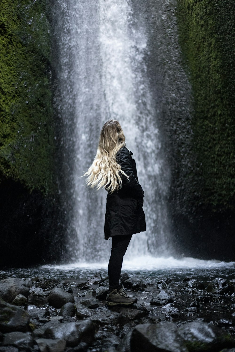 donna in cappotto nero in piedi su pietra davanti alle cascate