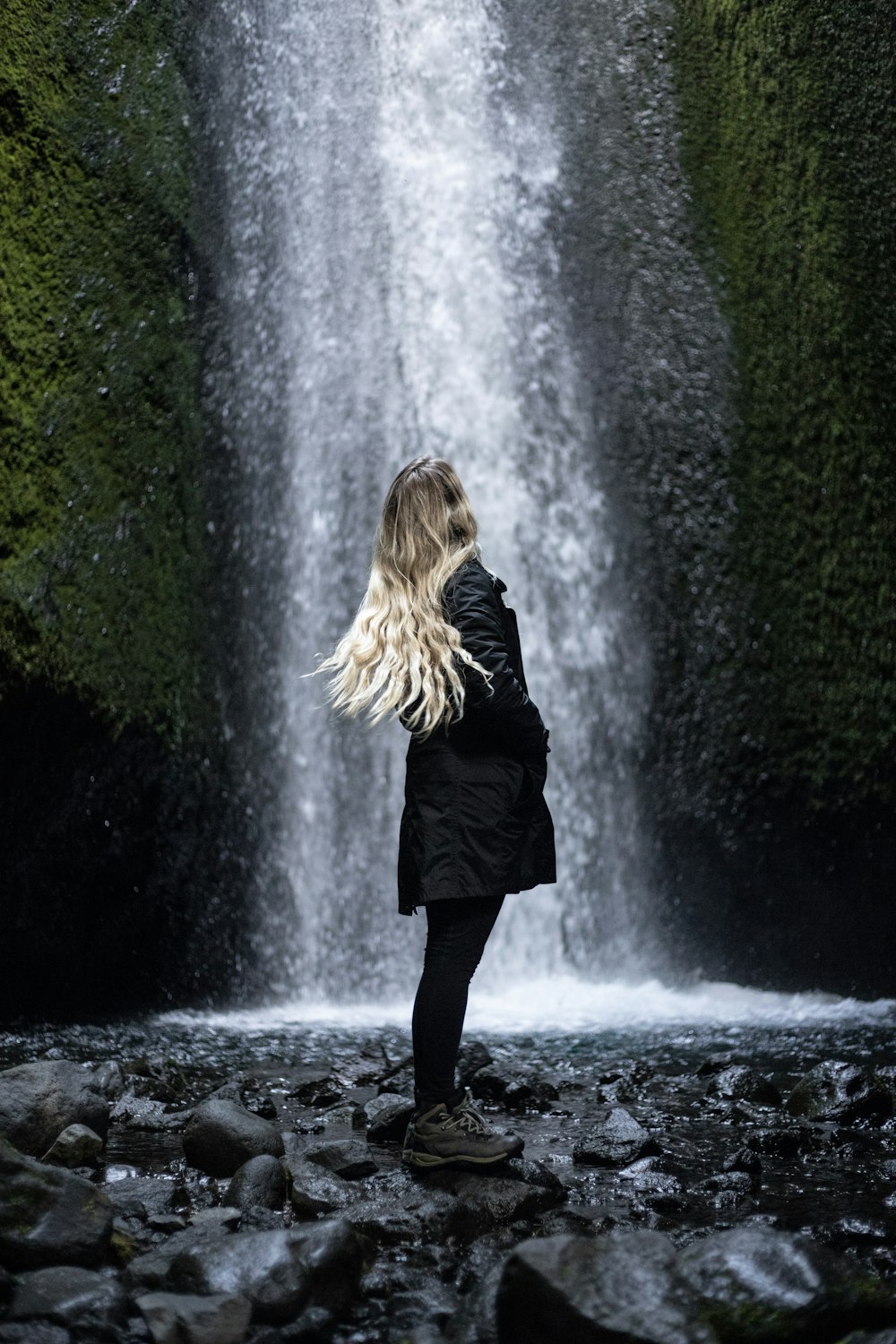 woman in black coat standing on stone in front of waterfalls