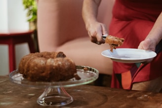 A person wearing a red apron slices a piece of cake onto a white plate, with a bundt cake resting on a glass cake stand on a wooden table. The background features parts of a pink chair and green foliage.