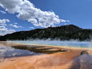 A vibrant hot spring with colorful mineral deposits and steaming water is set against a backdrop of evergreen forests and a blue sky filled with fluffy white clouds.