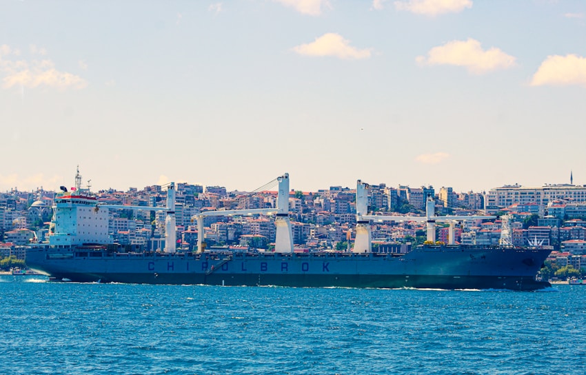 A large cargo ship is sailing in a body of water with a cityscape in the background, featuring numerous buildings stacked closely together. The ship has several large cranes and is labeled with the word CHINOLBROK. The sky is clear with a few clouds scattered throughout.