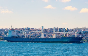 A large cargo ship is sailing in a body of water with a cityscape in the background, featuring numerous buildings stacked closely together. The ship has several large cranes and is labeled with the word CHINOLBROK. The sky is clear with a few clouds scattered throughout.