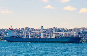 A large cargo ship is sailing in a body of water with a cityscape in the background, featuring numerous buildings stacked closely together. The ship has several large cranes and is labeled with the word CHINOLBROK. The sky is clear with a few clouds scattered throughout.