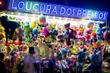 A vibrant and colorful carnival stall displays an array of plush toys and stuffed animals hanging from the ceiling. The illuminated sign above reads 'Loucura dos Prêmios', attracting people of all ages who are gathered around. The scene is lively with a variety of toys including bears, monkeys, and fantasy creatures.