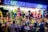 A vibrant and colorful carnival stall displays an array of plush toys and stuffed animals hanging from the ceiling. The illuminated sign above reads 'Loucura dos Prêmios', attracting people of all ages who are gathered around. The scene is lively with a variety of toys including bears, monkeys, and fantasy creatures.