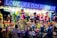 A vibrant and colorful carnival stall displays an array of plush toys and stuffed animals hanging from the ceiling. The illuminated sign above reads 'Loucura dos Prêmios', attracting people of all ages who are gathered around. The scene is lively with a variety of toys including bears, monkeys, and fantasy creatures.