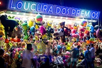 A vibrant and colorful carnival stall displays an array of plush toys and stuffed animals hanging from the ceiling. The illuminated sign above reads 'Loucura dos Pr&ecirc;mios', attracting people of all ages who are gathered around. The scene is lively with a variety of toys including bears, monkeys, and fantasy creatures.