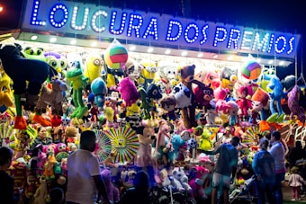 A vibrant and colorful carnival stall displays an array of plush toys and stuffed animals hanging from the ceiling. The illuminated sign above reads 'Loucura dos Prêmios', attracting people of all ages who are gathered around. The scene is lively with a variety of toys including bears, monkeys, and fantasy creatures.