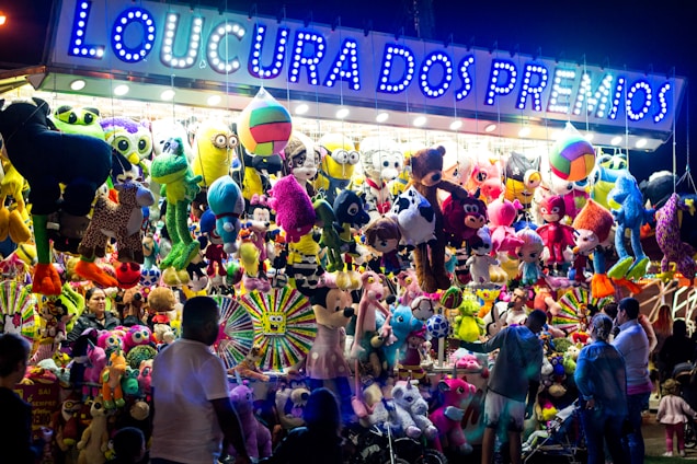 A vibrant and colorful carnival stall displays an array of plush toys and stuffed animals hanging from the ceiling. The illuminated sign above reads 'Loucura dos Pr&ecirc;mios', attracting people of all ages who are gathered around. The scene is lively with a variety of toys including bears, monkeys, and fantasy creatures.