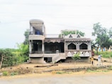 An unfinished two-story house made of concrete and bricks, featuring a cylindrical balcony on the left side and geometric patterns on the facade. The surrounding area has trees and bushes, with a dirt pathway in front. Two people are engaged in some activity on the ground outside the building.