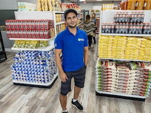 A man wearing a blue polo shirt and a cap stands in a well-organized store aisle. Shelves are stocked with various products, including coffee packets, rice bags, and beverages. Two women can be seen in the background shopping.