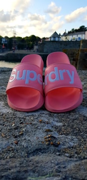 A pair of pink slide sandals with the word 'Superdry' printed in white on the straps are placed on a rough, textured surface. The background features a coastal scene with a calm waterfront, trees, and buildings under a partly cloudy sky.