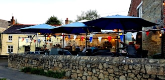 An outdoor restaurant or cafe with blue umbrellas over tables. There are several people seated, and string lights and colorful bunting decorate the space. The setting is adjacent to a stone wall and surrounded by a mix of old buildings.