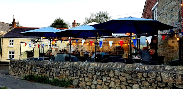 An outdoor restaurant or cafe with blue umbrellas over tables. There are several people seated, and string lights and colorful bunting decorate the space. The setting is adjacent to a stone wall and surrounded by a mix of old buildings.