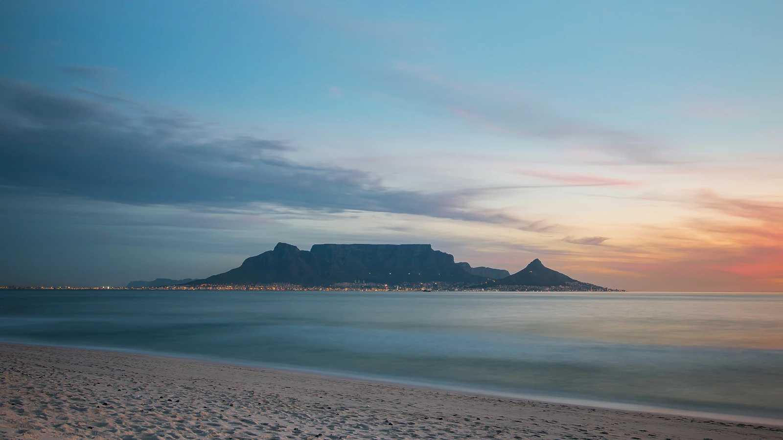 Table Mountain seen across the bay at dusk from a wide sandy beach, city lights glowing at its base