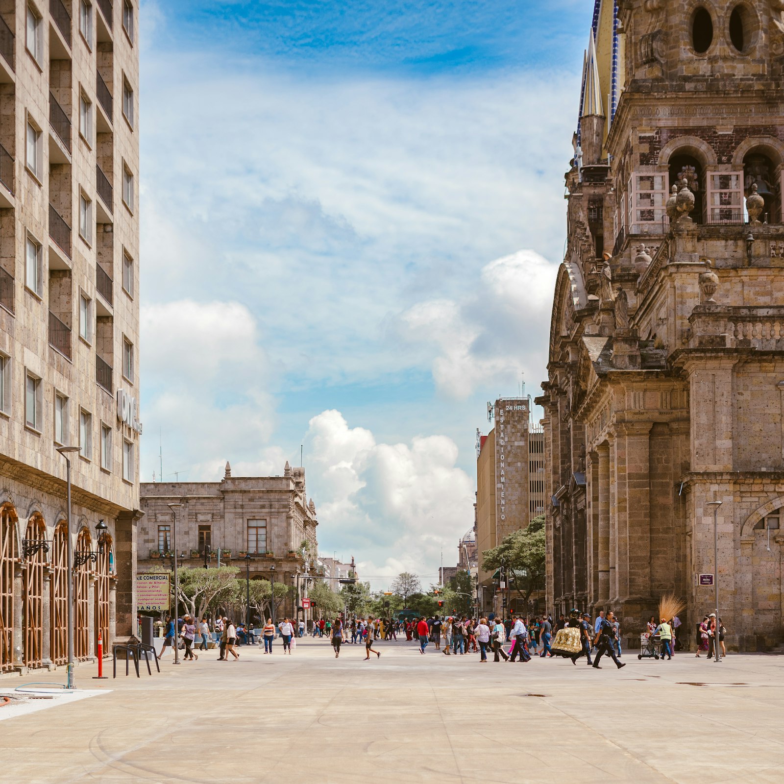 Pedestrians crossing a wide stone plaza beside the carved facade of Guadalajara's cathedral under a partly cloudy sky
