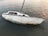 A weathered, white sailboat with visible peeling paint and patches of rust floats on calm water. The boat has a small cabin with dark windows and a visible number '70828' on its side. The mast is intact, though the sails are not present.