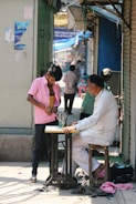 A man sits on a wooden stool working at a sewing machine on a narrow, busy street. Next to him stands a young boy wearing a pink shirt, trying on or adjusting clothes. The background shows several people walking and shops with various signs and colors.