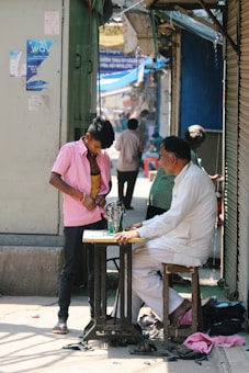 A man sits on a wooden stool working at a sewing machine on a narrow, busy street. Next to him stands a young boy wearing a pink shirt, trying on or adjusting clothes. The background shows several people walking and shops with various signs and colors.