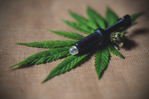A black object resembling a syringe or vape pen lies on top of a set of green cannabis leaves, with a cannabis bud beside it on a burlap fabric background.