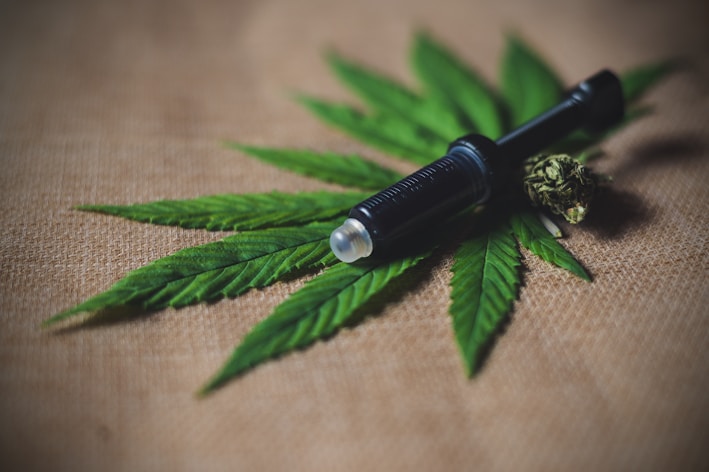 A black object resembling a syringe or vape pen lies on top of a set of green cannabis leaves, with a cannabis bud beside it on a burlap fabric background.