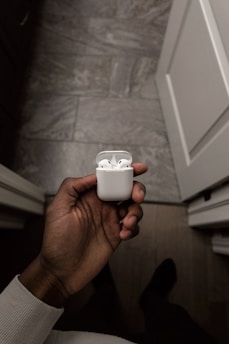 A hand is holding a white wireless earphones case against a modern tiled floor backdrop. The earphones are neatly packed inside the case.