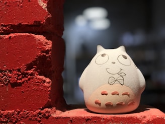 A ceramic figurine resembling a whimsical character is placed on a rough, red brick surface. The character has a cartoonish expression with large eyes, a bow tie, and small ears. The brick texture is prominent, contrasting with the smooth surface of the figurine.
