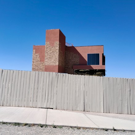 A modern building with a mix of stone and stucco facade stands behind a tall, light-colored wooden fence. The sky is clear and blue, and a concrete sidewalk runs parallel to the fence, with some sparse grass and gravel in the foreground.