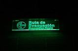 A glowing green illuminated evacuation route sign with text in both Spanish and English. The sign includes a left-pointing arrow and is mounted on a dark background.