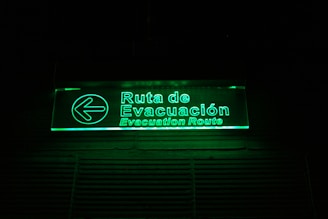A glowing green illuminated evacuation route sign with text in both Spanish and English. The sign includes a left-pointing arrow and is mounted on a dark background.