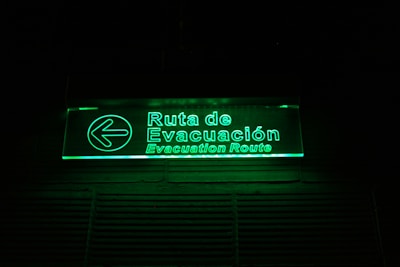 A glowing green illuminated evacuation route sign with text in both Spanish and English. The sign includes a left-pointing arrow and is mounted on a dark background.