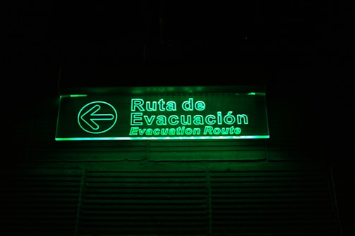 A glowing green illuminated evacuation route sign with text in both Spanish and English. The sign includes a left-pointing arrow and is mounted on a dark background.
