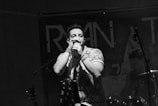 A male singer passionately performing with a microphone, wearing a patterned shirt. In the background, a drummer is visible, and there are musical instruments like a guitar and drum set. The setting appears to be a live music venue with a dark ambiance.