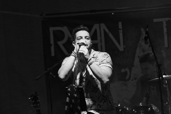 A male singer passionately performing with a microphone, wearing a patterned shirt. In the background, a drummer is visible, and there are musical instruments like a guitar and drum set. The setting appears to be a live music venue with a dark ambiance.
