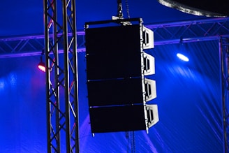 A set of large speakers is suspended from a metal truss structure. Vibrant lights in shades of blue and purple illuminate the scene, creating a dramatic effect. The setup appears to be part of a stage or concert venue, emphasizing professional sound equipment.