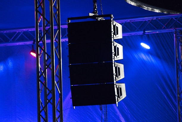A set of large speakers is suspended from a metal truss structure. Vibrant lights in shades of blue and purple illuminate the scene, creating a dramatic effect. The setup appears to be part of a stage or concert venue, emphasizing professional sound equipment.