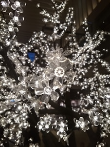 A close-up of artificial flowers made from clear plastic, illuminated by small LED lights, creating a shimmering effect. The branches appear densely covered with these glowing blossoms set against a dark background.